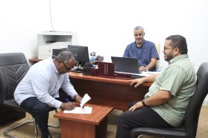 WhatsApp Image 2026-07-06 at 9.50.42 PM (2) - جامعة سبها | Sebha University Three men in an office: one signs documents at a small table while two others watch nearby in a meeting room.