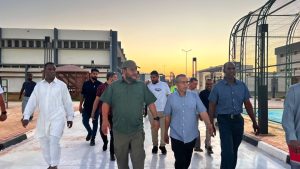 WhatsApp Image 2026-07-09 at 10.21.17 AM - جامعة سبها | Sebha University Group of officials walking outdoors at sunset on a site visit, past a high-security fence and industrial buildings.