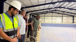 Team of inspectors standing along the edge of an empty indoor pool during a site visit; foreground man in hard hat and high-visibility vest with a lanyard. (informative)