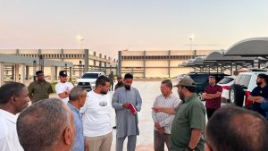 A group of men standing in a parking area, one person in traditional grey clothing showing a red notebook to others as they listen nearby at sunset.