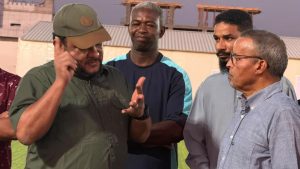 WhatsApp Image 2026-07-09 at 10.21.18 AM - جامعة سبها | Sebha University Man in a green shirt and olive cap speaks to a group of men on a grassy outdoor area, gesturing with his hands.