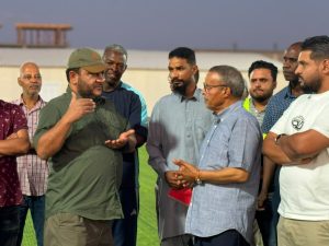 WhatsApp Image 2026-07-09 at 10.21.19 AM (1) - جامعة سبها | Sebha University Group of men on a grassy field engaged in discussion, two foreground men gesturing as others listen.