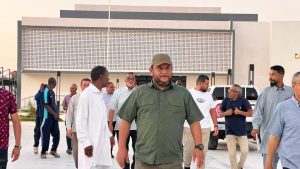 Group of men walking and standing in a parking lot outside a modern building, center foreground man in green shirt and cap looking ahead.