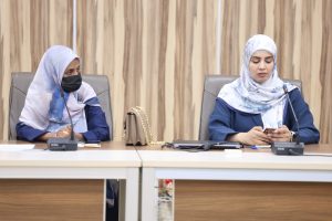 Two women in hijabs sit at a conference table with microphones; one wears a black mask and looks left, the other looks at a phone in front of a beige curtain backdrop