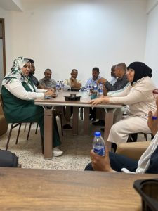 WhatsApp Image 2026-07-12 at 11.32.21 PM (2) - جامعة سبها | Sebha University Group of adults seated around a rectangular meeting table, discussing with water bottles on the table in a small conference room.