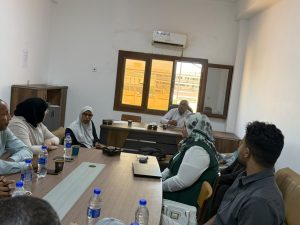 WhatsApp Image 2026-07-12 at 11.32.22 PM (1) - جامعة سبها | Sebha University Meeting in progress: several coworkers seated around a wooden conference table with water bottles and cups in a bright office.