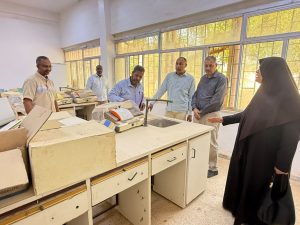WhatsApp Image 2026-07-12 at 11.32.23 PM (1) - جامعة سبها | Sebha University Woman in a black abaya speaks to a group of men in a laboratory or classroom, gesturing toward dusty lab benches with vintage weighing scales near large windows.