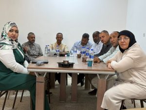 WhatsApp Image 2026-07-12 at 11.32.24 PM (1) - جامعة سبها | Sebha University Group of people seated around a rectangular meeting table in a bright room, with water bottles and cups on the table, appearing to discuss something thoughtfully.