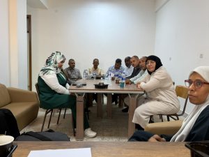 WhatsApp Image 2026-07-12 at 11.32.25 PM - جامعة سبها | Sebha University Group of adults seated around a rectangular table in a plain meeting room, discussing with water bottles on the table.