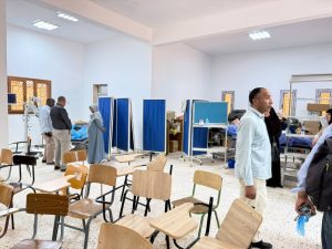 WhatsApp Image 2026-07-12 at 11.32.26 PM (1) - جامعة سبها | Sebha University A clinic or waiting area with blue privacy screens, empty chairs, and several people standing and talking near examination beds.