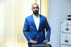 WhatsApp Image 2026-07-14 at 1.26.50 PM - Copy - جامعة سبها | Sebha University Man in a blue suit standing beside a chair in an office, beige vertical blinds behind him and a safe visible on the right.