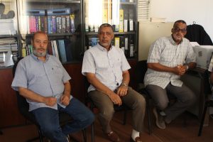 Three middle-aged men in button-down shirts seated in an office, with shelves of binders behind them.