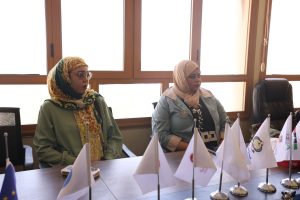 Two women wearing headscarves sit at a conference table with small flags in front of them, in a bright room with large wooden-framed windows behind.