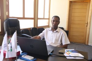 WhatsApp Image 2026-07-14 at 12.29.35 PM - جامعة سبها | Sebha University Man in a light shirt sits at a desk with an HP laptop in a sunlit office, wooden windows and a tall wooden door behind him.