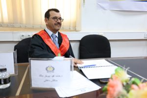 Man in academic gown with a red sash sits at a desk with papers and a bouquet in foreground, during a formal ceremony.