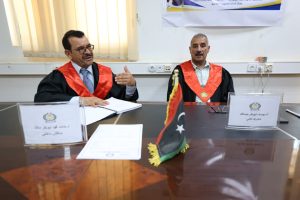 WhatsApp Image 2026-07-18 at 11.24.33 AM - جامعة سبها | Sebha University Two men in black judicial robes with red sashes sit at a conference table, a Libyan flag between them and documents in front of them.