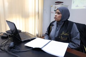 Woman in a hijab sits at a desk with an open laptop and a spiral notebook, focused on the screen nearby.