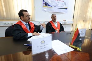 Two men in academic gowns with red sashes sit at a conference table, one speaking while the other looks on, in a formal ceremony setting with a flag on the desk and a poster on the wall behind them, suggesting a signing or presentation event.
