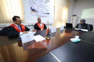 Three panelists sit at a polished conference table wearing red academic robes; a Libyan flag and banner are visible in the background.