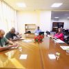 People seated around a long conference table in a brightly lit office, engaged in a meeting with papers and laptops.