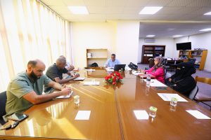 People seated around a long conference table in a brightly lit office, engaged in a meeting with papers and laptops.