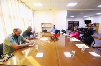 People seated around a long conference table in a brightly lit office, engaged in a meeting with papers and laptops.