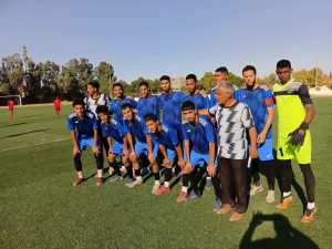 Group of young soccer players in blue uniforms posing for a team photo on a sunny outdoor field; goalkeeper in neon, black gloves on the right, and a coach in gray nearby.