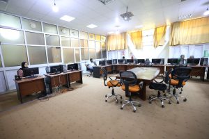 Office computer lab with multiple desks and monitor stations along the windows, people seated at computers working or chatting.