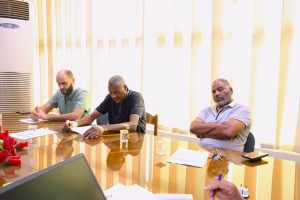 52190C02-D512-47F0-A3F6-A99DB154B7B8 - جامعة سبها | Sebha University Three men sit at a glass conference table reviewing documents in a bright room.
