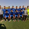 Eleven soccer players in blue jerseys pose for a team photo on a sunny field, with a neon-yellow goalkeeper on the right.