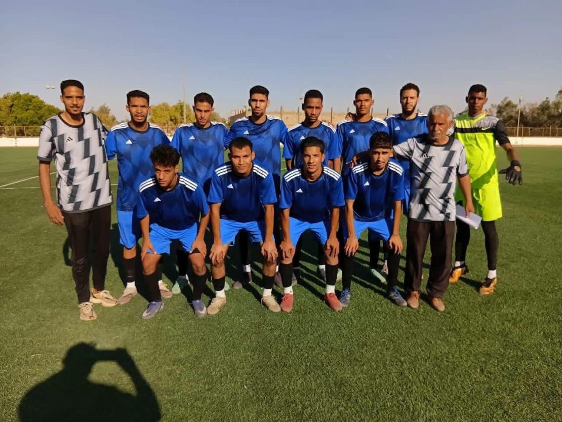 Eleven soccer players in blue jerseys pose for a team photo on a sunny field, with a neon-yellow goalkeeper on the right.