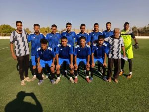 5AD05B9C-478C-4602-8B73-DCF749A3A758 - جامعة سبها | Sebha University Eleven soccer players in blue jerseys pose for a team photo on a sunny field, with a neon-yellow goalkeeper on the right.