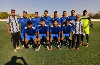 Eleven soccer players in blue jerseys pose for a team photo on a sunny field, with a neon-yellow goalkeeper on the right.