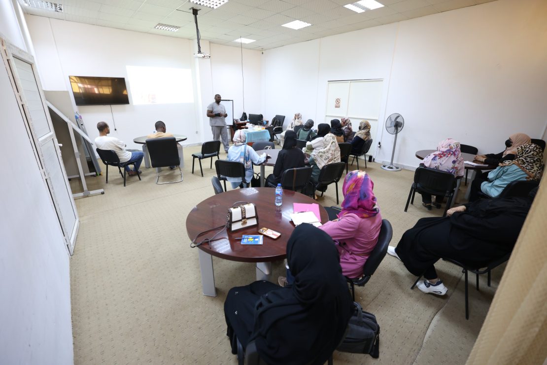 Instructor speaks to a classroom of adults seated in a semicircle, projection screen at front, white walls, and tables with notebooks and bottles nearby.