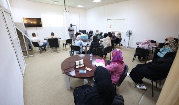 Instructor speaks to a classroom of adults seated in a semicircle, projection screen at front, white walls, and tables with notebooks and bottles nearby.
