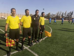 80A068A4-5CA9-4EDD-89AD-9B2FA4183C00 - جامعة سبها | Sebha University Four referees in yellow shirts stand on a soccer field near the sideline, with blue-clad players visible in the background.