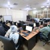 Computer lab with multiple people seated at desks, each with a monitor, keyboard, and mouse; a projector screen at the front of the room.