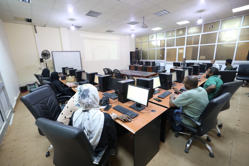 Computer lab with multiple people seated at desks, each with a monitor, keyboard, and mouse; a projector screen at the front of the room.