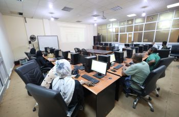 Computer lab with multiple people seated at desks, each with a monitor, keyboard, and mouse; a projector screen at the front of the room.