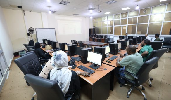 897C32DE-D785-4659-9568-DB17BDEEFFF1 - جامعة سبها | Sebha University Computer lab with multiple people seated at desks, each with a monitor, keyboard, and mouse; a projector screen at the front of the room.