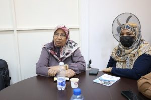 - جامعة سبها | Sebha University Two women in hijabs sit at a conference table during a meeting; one wears a black face mask and the table has water bottles and papers nearby.