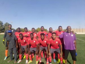 CB2387E5-4F78-4527-B41C-04969692FFB5 - جامعة سبها | Sebha University Soccer team in red shirts and gray shorts posing for a group photo on a sunny outdoor field with two coaches nearby.