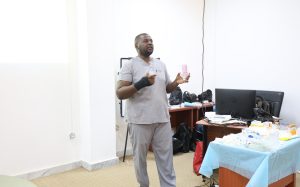 A man in gray medical scrubs speaks, holding a bottle in one hand and a small object in the other, standing beside a cluttered desk with supplies and a computer monitor in the background.