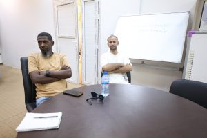 D6FE0308-9498-4DAE-BB51-1E9A9F872C5D - جامعة سبها | Sebha University Two men sit at a conference table in a meeting room, arms crossed, with a whiteboard behind them and a water bottle on the table.