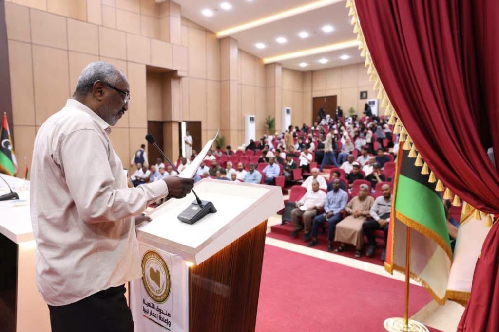 DBD293E7-5F24-4445-8E34-0CE8996EE22E - جامعة سبها | Sebha University Older man speaking at a podium in a large conference hall, addressing a seated audience.
