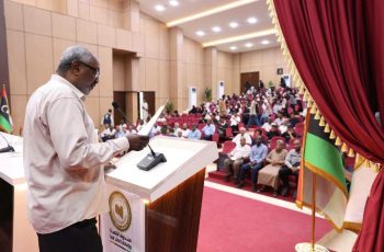 Older man speaking at a podium in a large conference hall, addressing a seated audience.