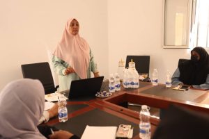 Woman in a pink hijab stands at a conference table giving a presentation to seated colleagues. A laptop and bottled water are on the table.