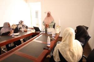 Six women wearing hijabs sit around a U-shaped conference table; a presenter in a light pink hijab stands at the front with a laptop nearby and bottled water on the table.