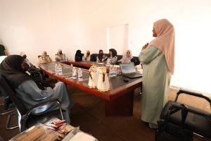 Women wearing hijabs sit around a conference table as a presenter stands at the front with a laptop nearby in a bright room.