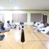 Six men sit around a long wooden conference table in a meeting room, with laptops and small flags at the center of the table.
